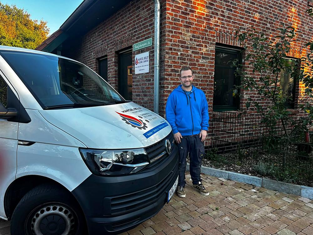 Marcel Boldt vor seinem Firmenfahrzeug am Betriebssitz, daneben das Firmenschild an der Hauswand
