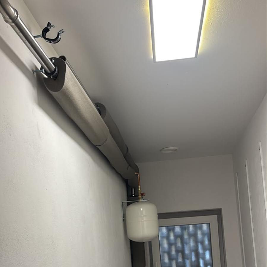 Gedämmte Heizungsleitungen mit Rohrschellen unter der Decke eines Flurs, darunter ein weißes Ausdehnungsgefäß